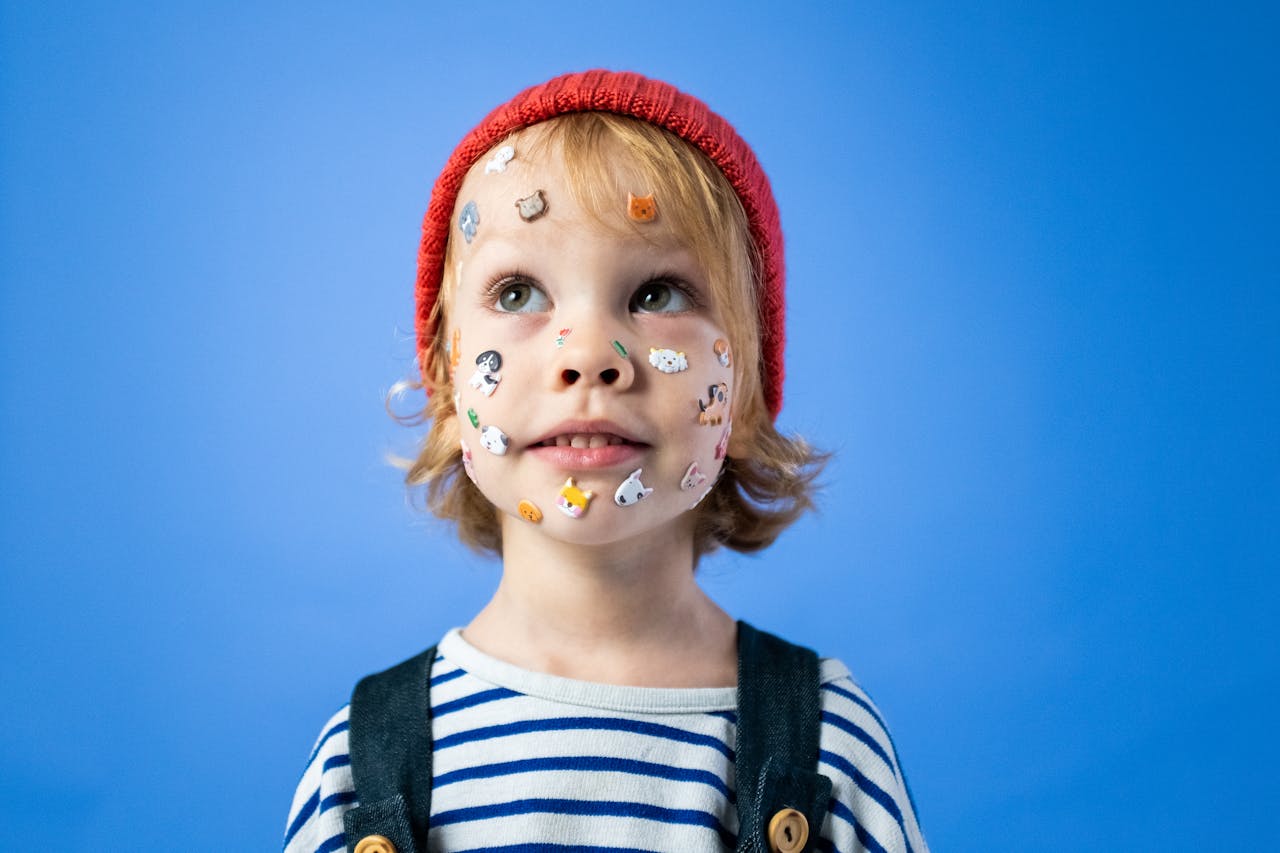 Playful portrait of a young child wearing a red hat with face stickers and a striped shirt against a blue background.